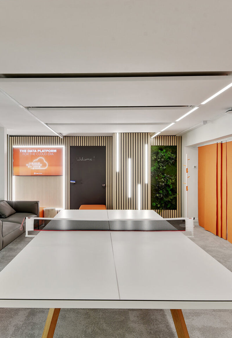Modern office space featuring a white ping pong table at the center, with a blackboard door labeled 'Welcome!', vertical wood paneling, LED light strips, a green plant wall, an orange digital display about a cloud data platform, a gray couch on the left, and orange curtains on the right.